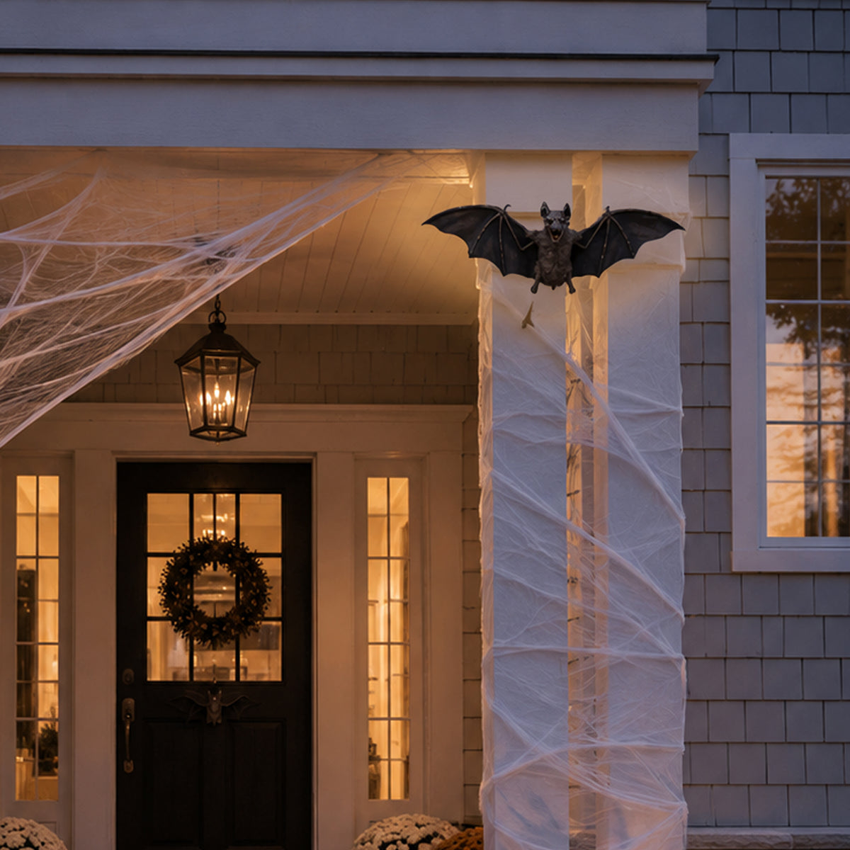 A realistic flying bat Halloween decoration hanging from a porch column, with white cobwebbing draped across the front porch and a lit lantern above a decorated door.