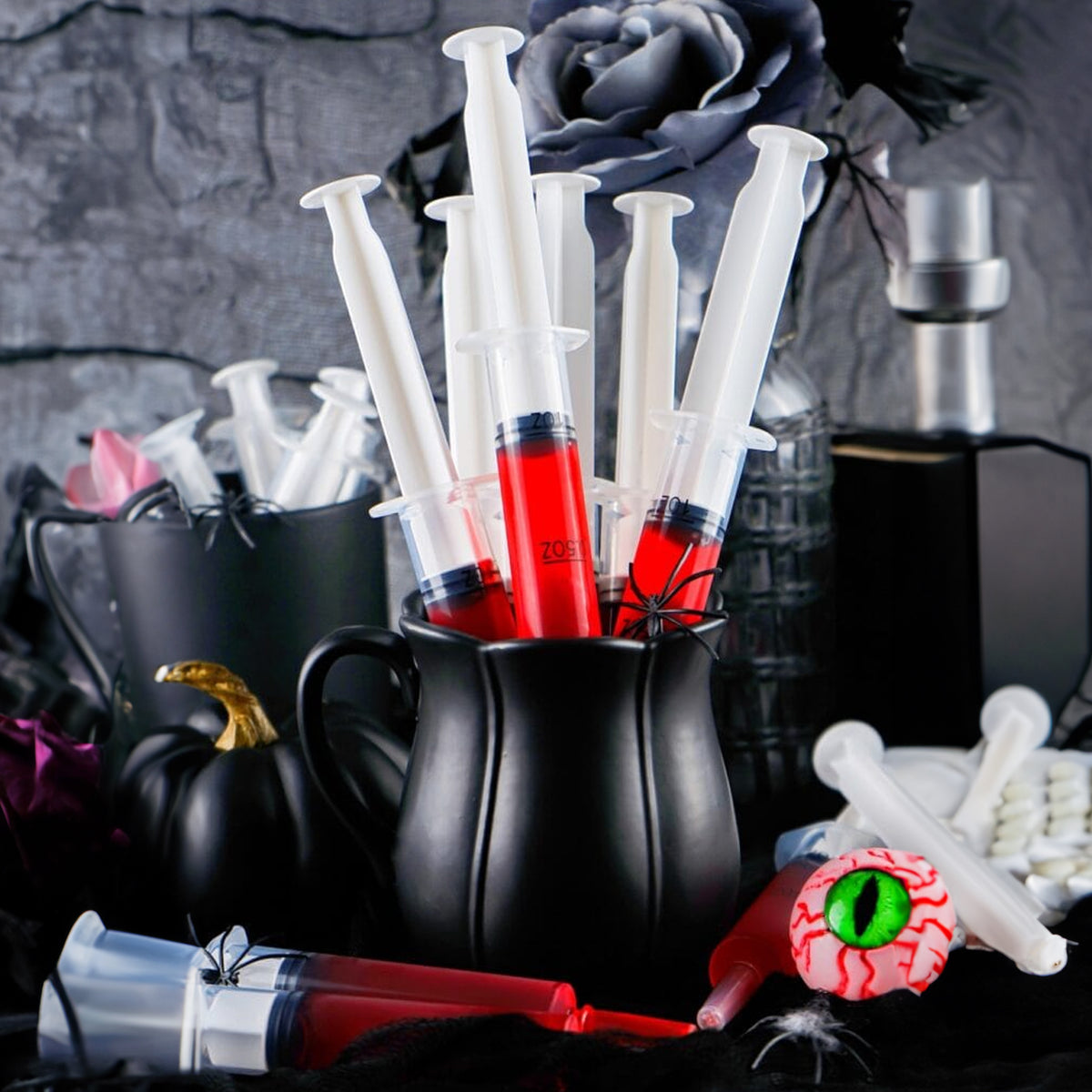 Black plastic syringe cup holders filled with red liquid, styled with Halloween props including a small black pumpkin, a eyeball decoration, and a dark rose against a gray stone-textured background.
