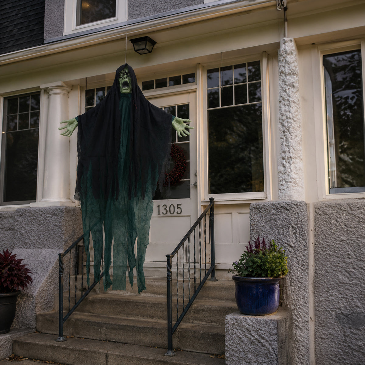 A spooky Halloween decoration of a ghostly figure draped in black fabric stands on a front porch with its arms outstretched. The figure has green skin and a tattered dark green body, set against a house entrance with stone steps and columns.