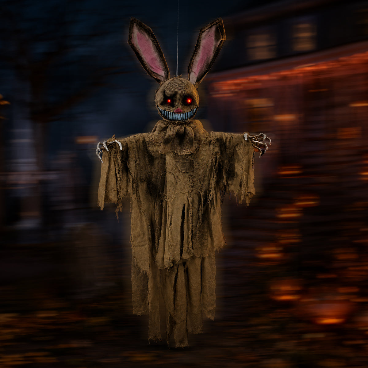 A creepy hanging bunny ghost decoration with long ears, a tattered brown rag body, skeletal hands, and glowing red and blue eyes, set against a blurred dark street background at night.