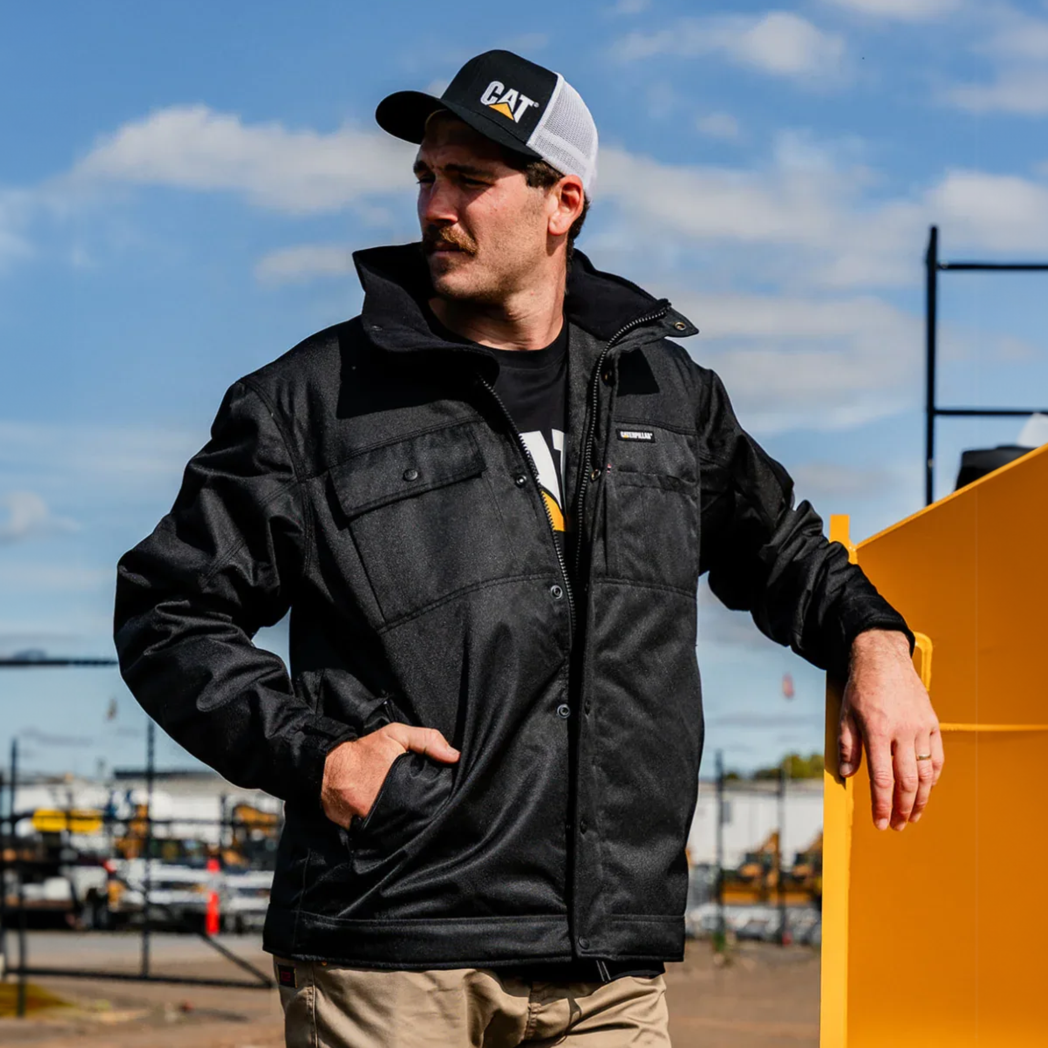 A man wearing a black CAT-branded work jacket, black and white CAT trucker hat, and khaki pants, standing outdoors next to a large yellow machine under a blue sky.