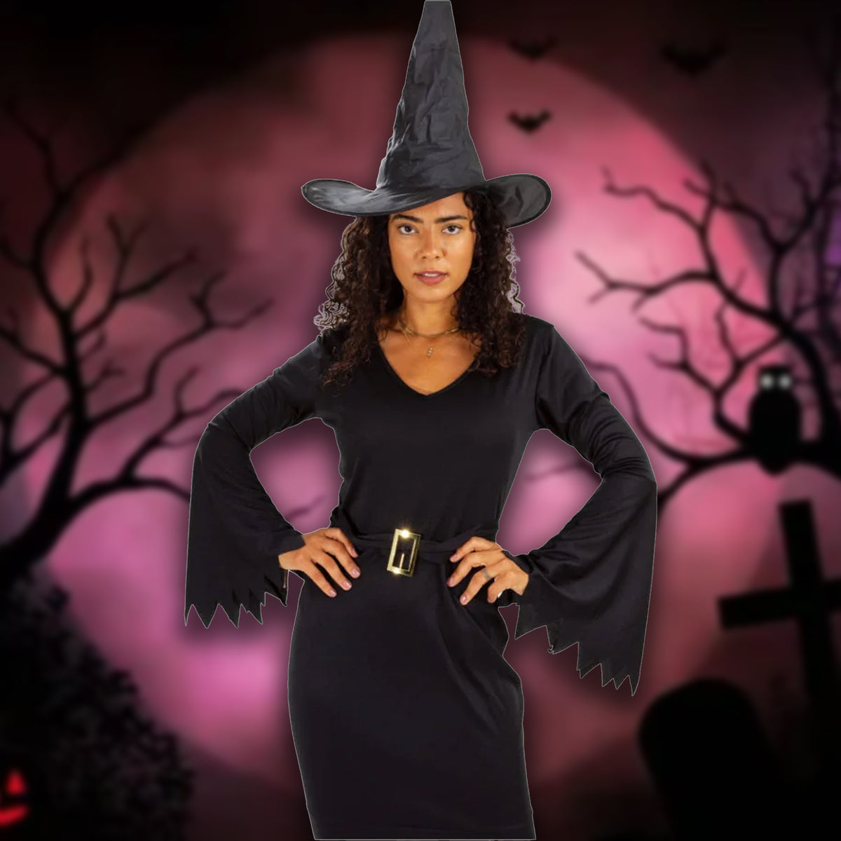 A woman wearing a black witch costume with a pointed hat, long flared sleeves, and a gold belt buckle, posed against a spooky Halloween background with a pink moon, silhouetted trees, and a cross.