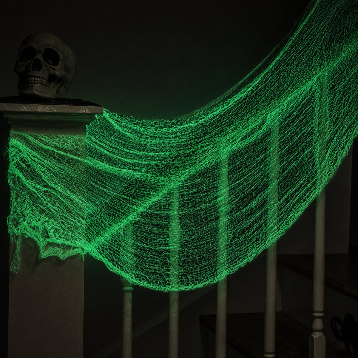 Glowing green cobweb Halloween netting draped over a staircase railing, with a skull decoration on the left side in a dark room.