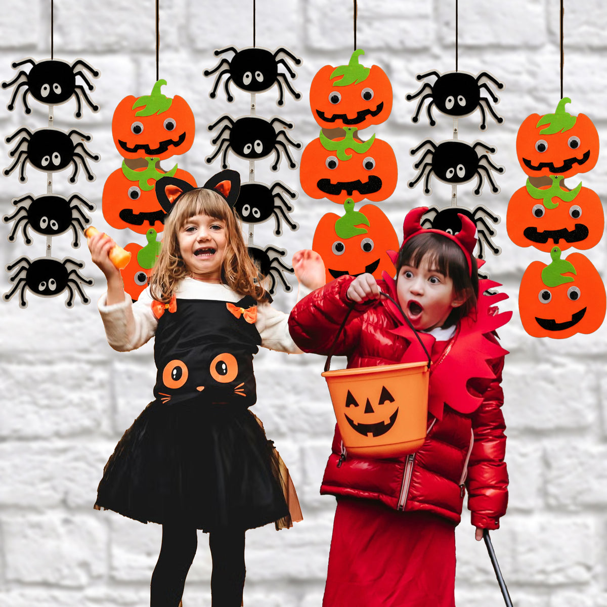 Two children in Halloween costumes pose in front of a white brick wall decorated with hanging orange jack-o'-lanterns and black spiders. One child wears a black cat costume with orange eyes, and the other wears a red costume and holds an orange pumpkin bucket.