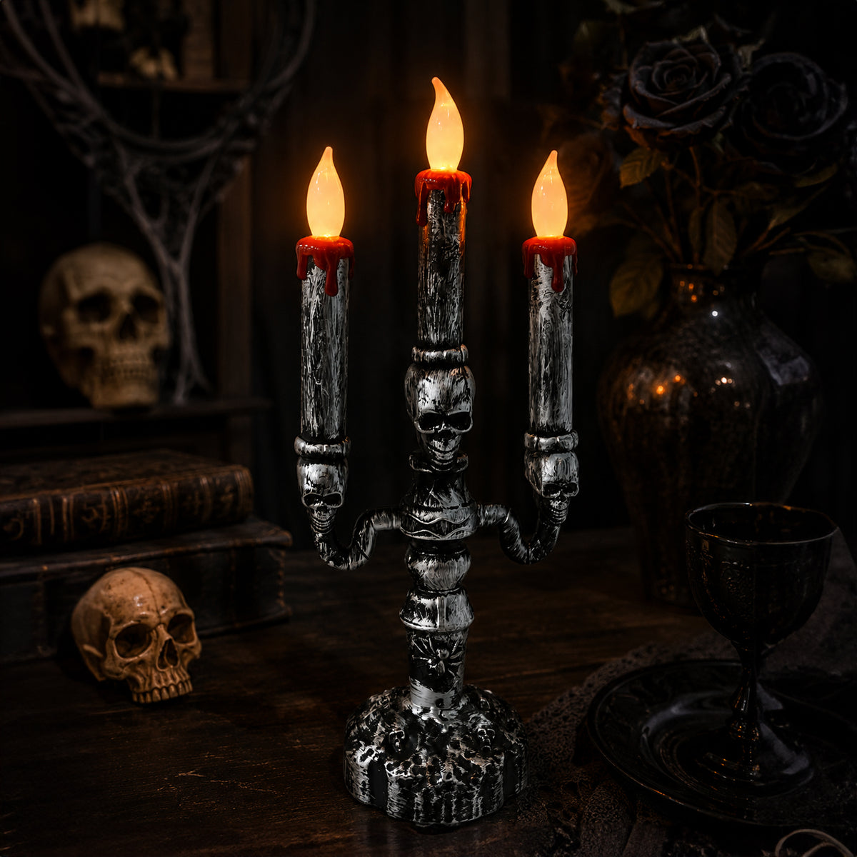 A gothic-style three-arm candelabra candle holder with a skull motif, holding three lit faux candles with red wax details, set on a dark wooden table with skulls, books, and a goblet in a moody room.